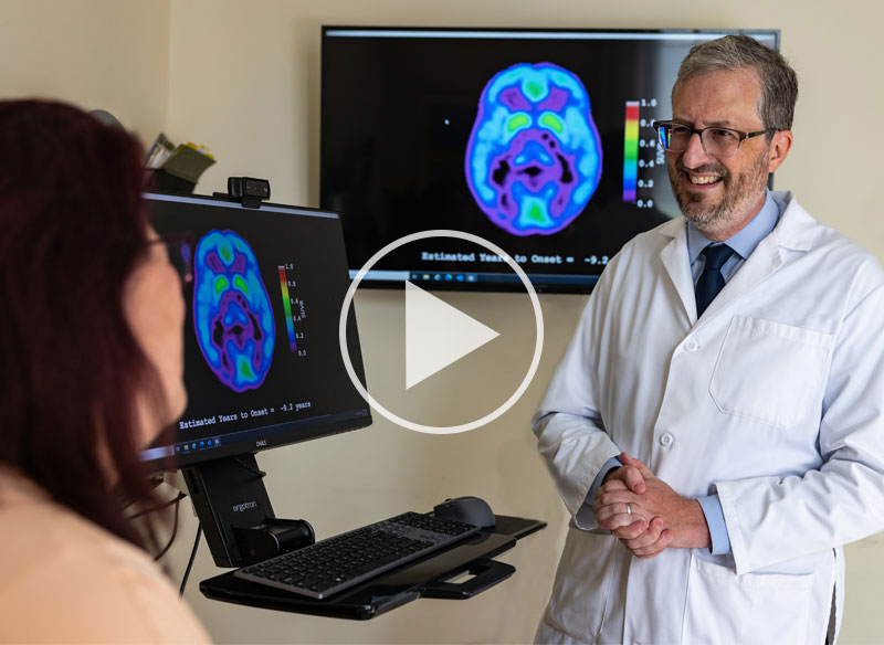 Ted Huey speaking with patient with brain scan images on monitors in the room. Play button superimposed over the image