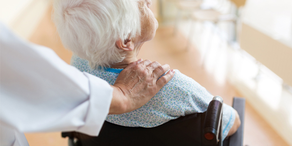 An elderly patient in a wheelchair, seen from behind, with a reassuring physician's hand on their shoulder