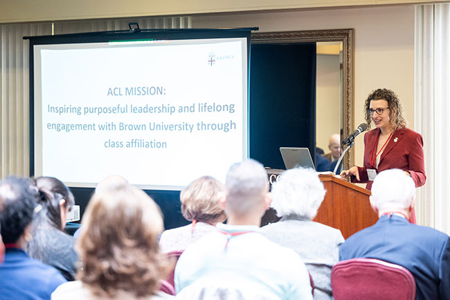 ACL Board President Sarah Gomel speaking into a microphone with meeting attendees in foreground